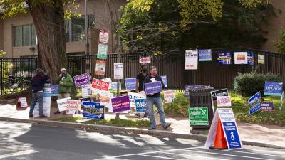 A person holds a sign while sanding among other candidate signs on a sidewalk corner