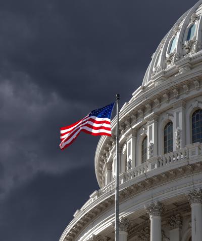 A close up of the dome of the U.S. Capitol Building with an American flag flying and a dark cloudy sky in the background.